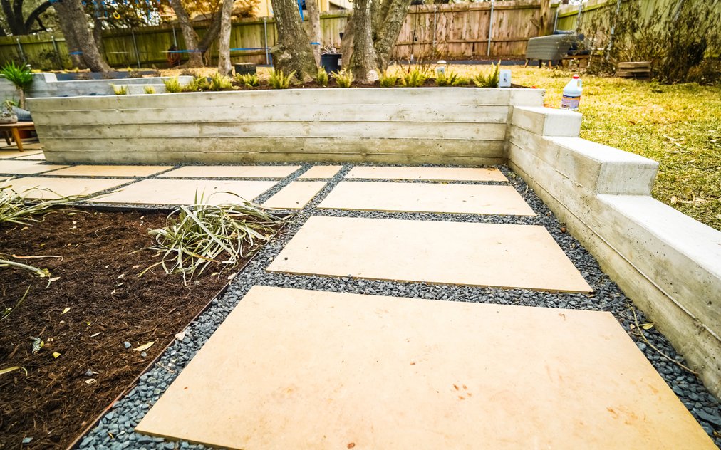 Board-formed concrete retaining wall with built-in steps holding back a raised lawn above a large-format paver and gravel patio in an Austin backyard near Shoal Creek